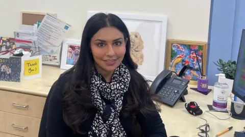 df0146c0-9cf8-11ef-9d47-3962c94c3bbb-jpg Deepali Misra-Sharp Dr Deepali Misra-Sharp sits at her work desk, with pictures and cards behind her.